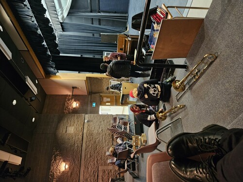 Several students and a instructor in a high school theatre. Two students in the front of the picture have opened two trombone cases and the trombones are sitting behind them. Two students meet with the instructor in middle of the photo. Another student