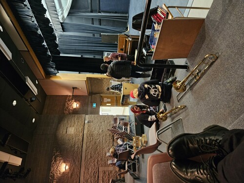 Several students and a instructor in a high school theatre.  Two students in the front of the picture have opened two trombone cases and the trombones are sitting behind them. Two students meet with the instructor in middle of the photo.  Another student 