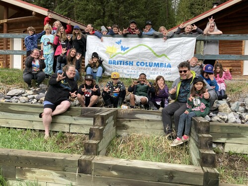 A group of students and some adults sit and stand around a deck and stairs with a large sign that says BC Provincial Outreach Program for Deaf and Hard of Hearing students
