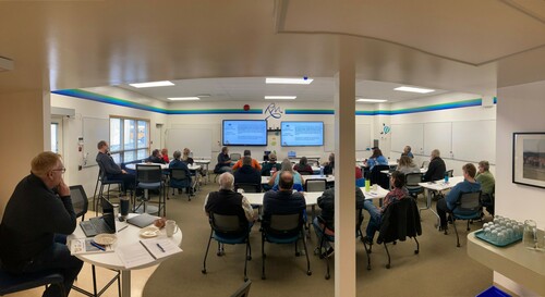 A wide angle photo from the back of a white room. A group of adults sitting at tables and are facing two tv screens.