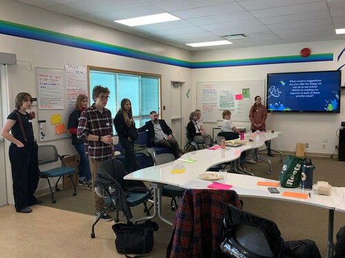 Students and teachers listening to a speaker. There are white boards with papers and text on them.