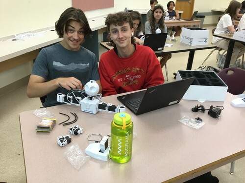 Two students are sitting on at a desk. The student on the left has ear-length hair, is smiling and is wearing a grey t-shirt. The student on the right has a mullet, has an airpod in their right ear, is smiling and is wearing a red sweatshirt. There is a