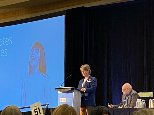 Middle aged woman at a podium with a screen behind her.  The screed says candidate speeches with a young female child with a big open mouthed smile