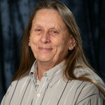 Senior aged woman with grey brown long hair.  Wearing a beige collared shirt with white and grey vertical stripes