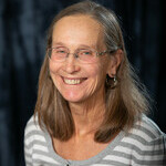 Senior aged woman with grey brown collarbone length hair smiling wearing a grey and white striped shirt