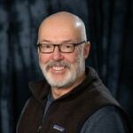 Middle aged bald man wearing glasses in black vest with blue shirt on underneath