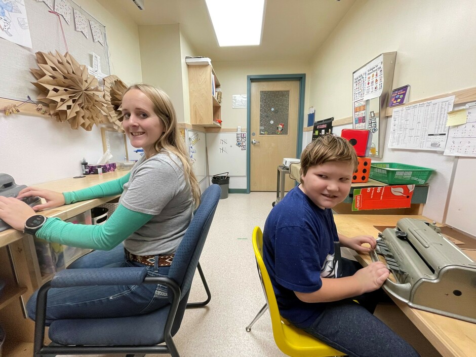 A young woman with long blonde hair and a young boy with short brown hair smile at the camera. She is wearing jeans with a grey t-shirt with green sleeves and jeans. He is wearing a blue t-shirt with jeans. Both are sitting in a small office at Perkins