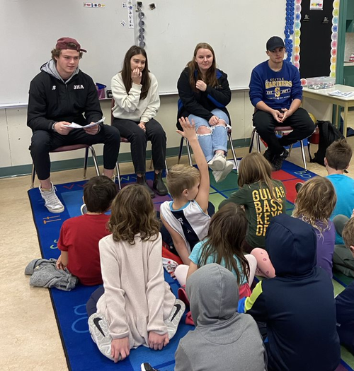 Four teenages face a group of elementary students. One child has their hand up to ask a question.