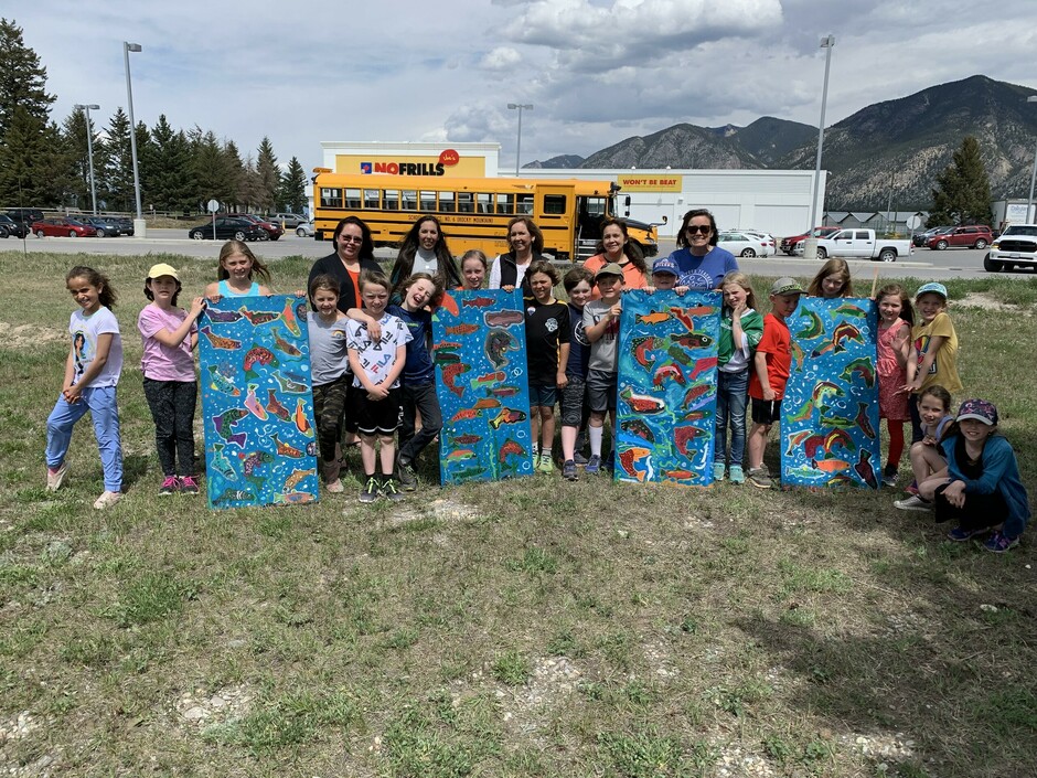 A class of students holding four blue panels with salmon pained on them. Bus parked behind them.